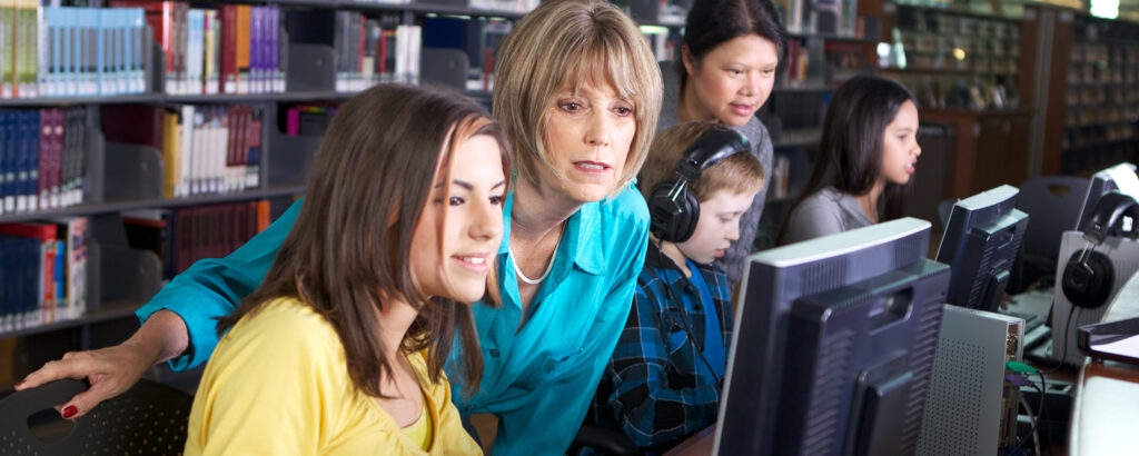 Educators helping students using desktop computers in library.