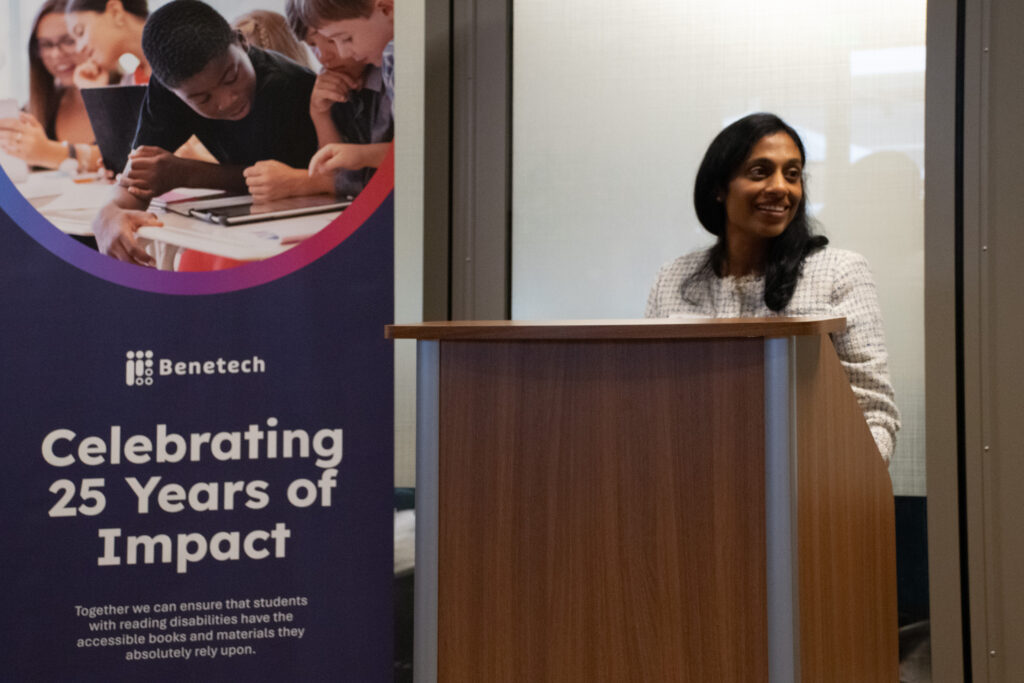 Pallavi Gondipalli smiles at the podium during a Benetech 25th anniversary celebration, with a branded banner beside her.